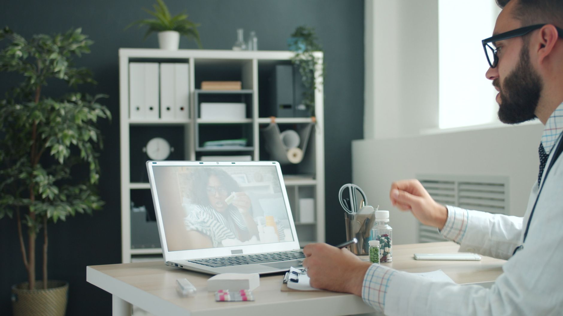Man with glasses and beard in white shirt working at desk with laptop in modern office with shelving and plants