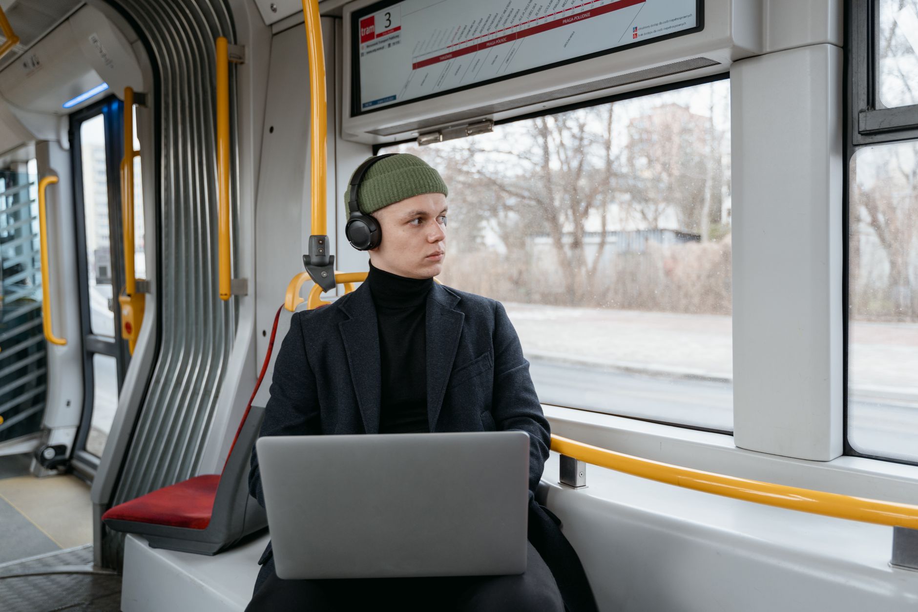 Person on public transit bus using laptop while wearing headphones and green beanie