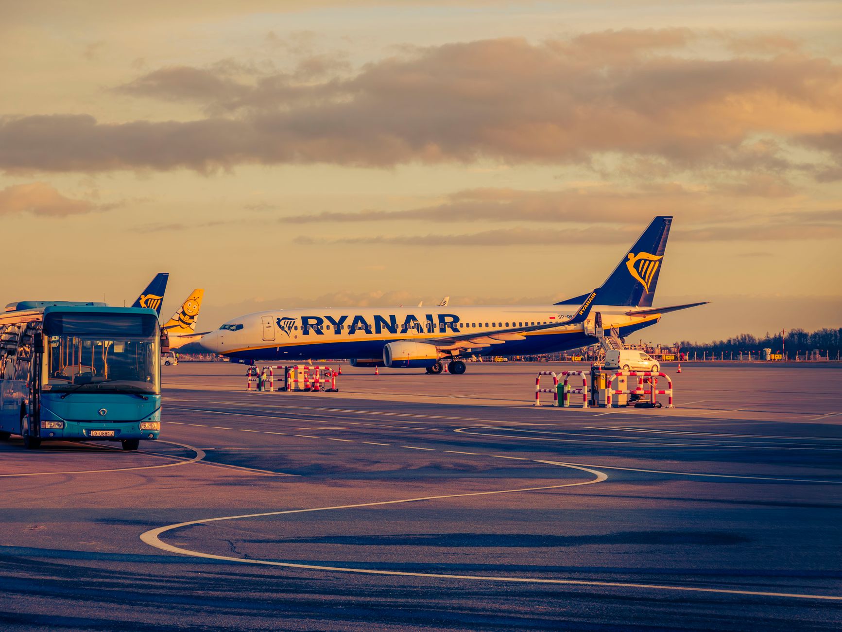 Blåt og gult fly parkeret på lufthavnslandingsbane med blå shuttle-bus i forgrunden ved solnedgang.
