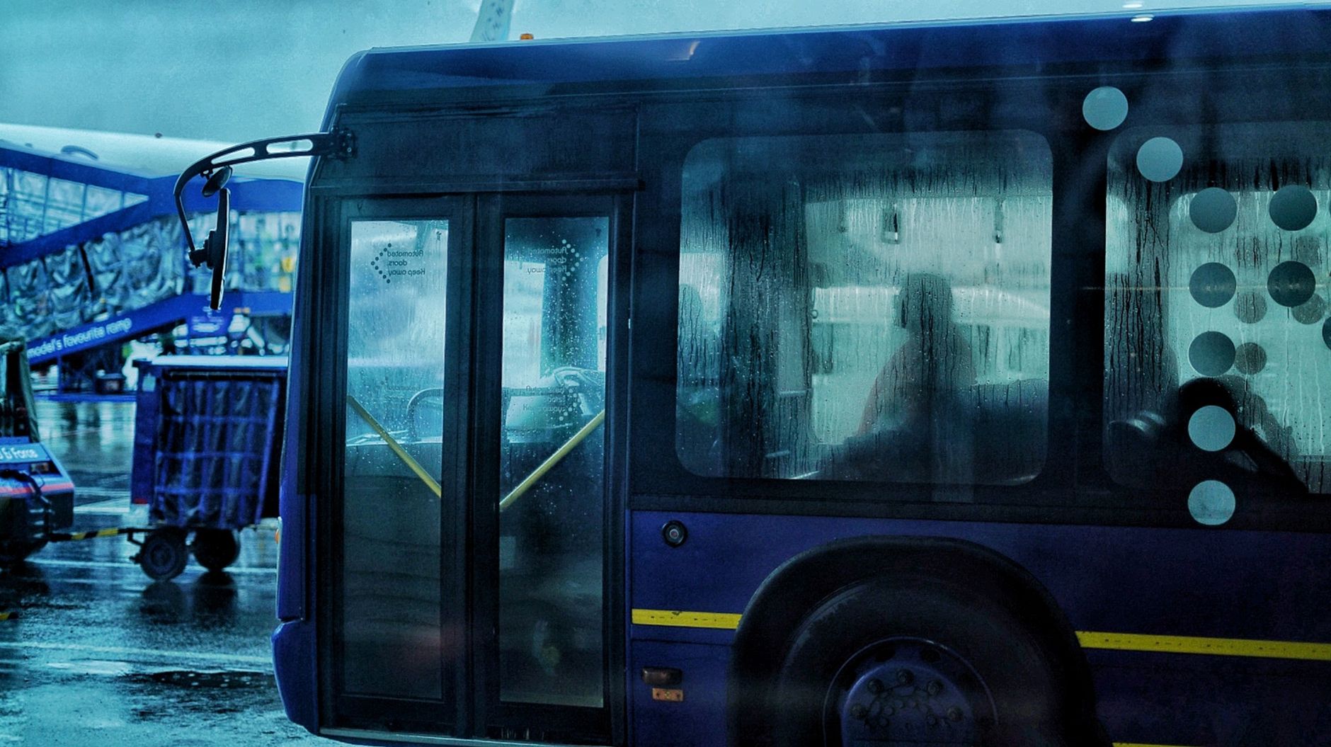 Blå shuttle-bus med våde vinduer og dørे på regnet lufthavnsterminal.